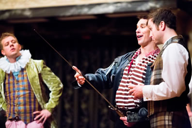 Bobadill in red striped shirt and black leather jacket, Ed Knowell in brown tartan vest and pants with white shirt, and Master Stephen to the left, hands on hips, wearing purple and gold checked vest with blue buttons, blue shirt, green felt jacket, pink pants, and Elizabethan ruff collar